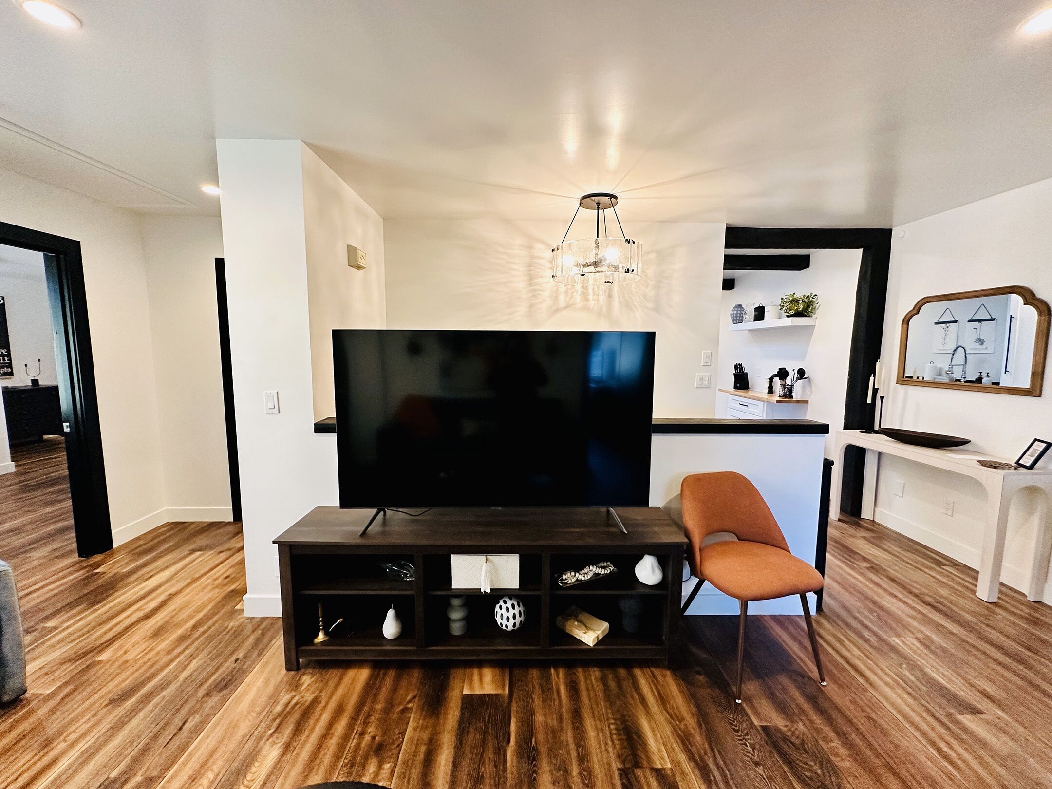 A modern living room with a flat-screen TV, a dark wooden entertainment unit, a brown leather chair, and a kitchen area in the background.
