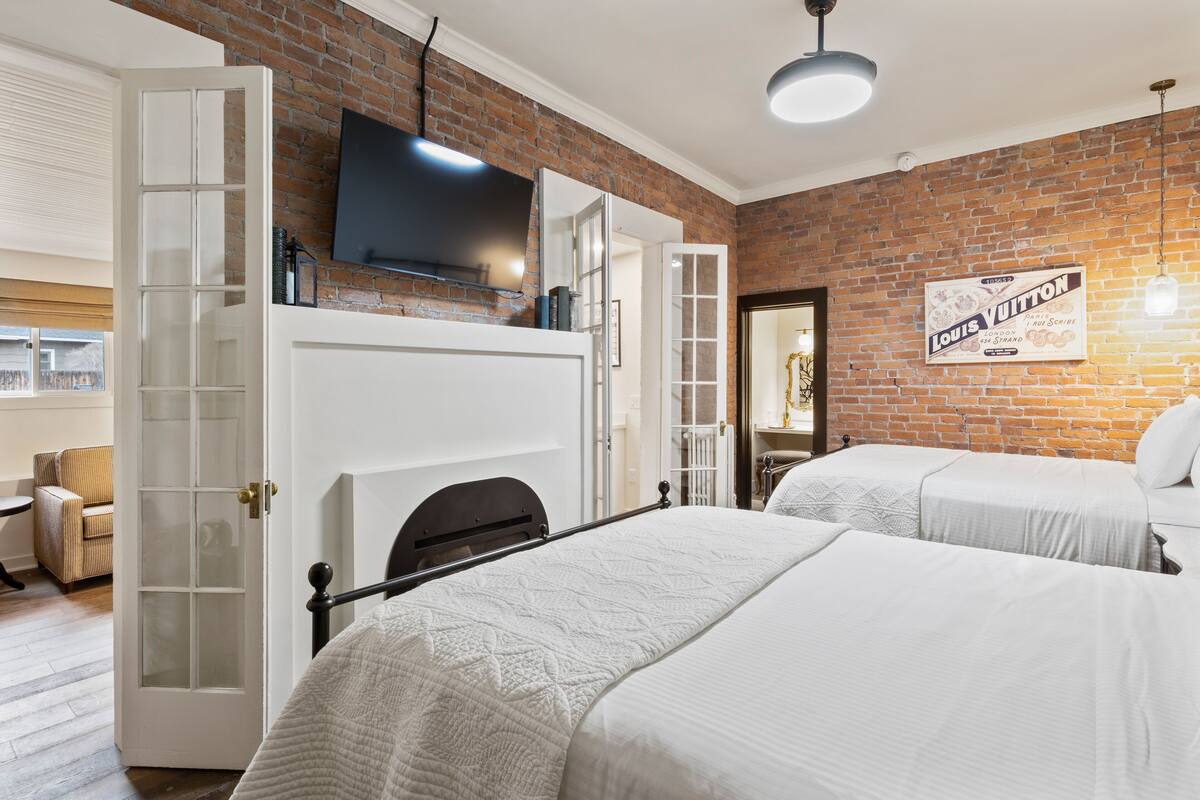 A hotel room with a brick wall, a flat-screen TV, a Louis Vuitton sign, and two beds with white bedding.