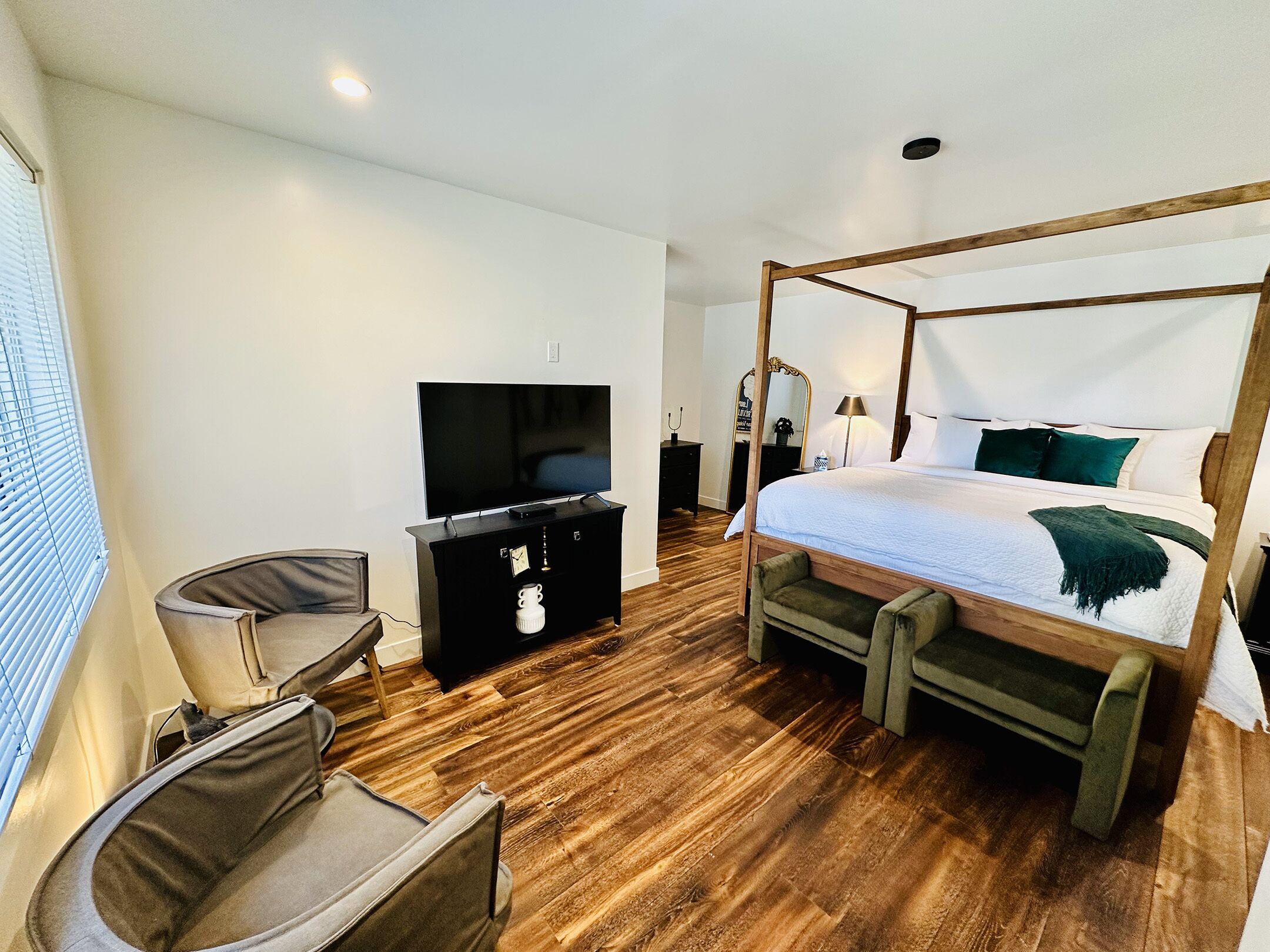 A modern hotel room with a four-poster bed, a flat-screen TV, a wooden dresser, and a comfortable armchair.