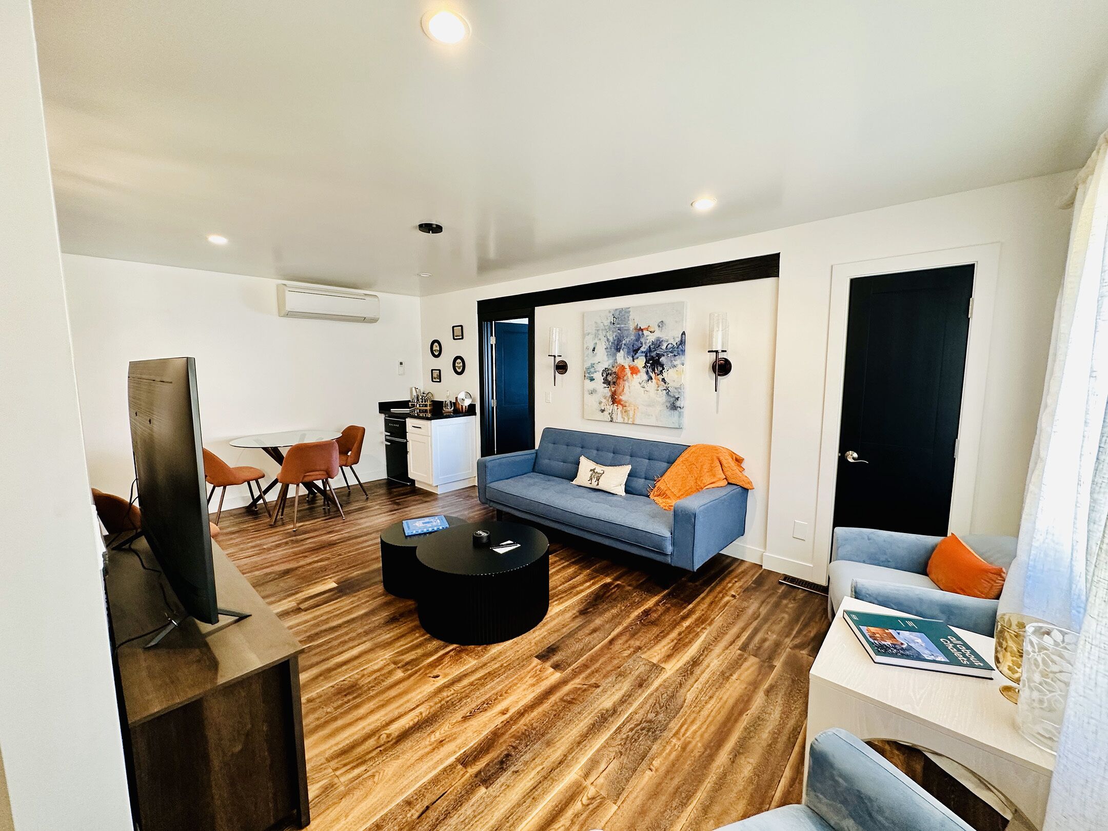 A modern living room with a blue sofa, a coffee table, and a TV. There is a kitchen area with a dining table and chairs.