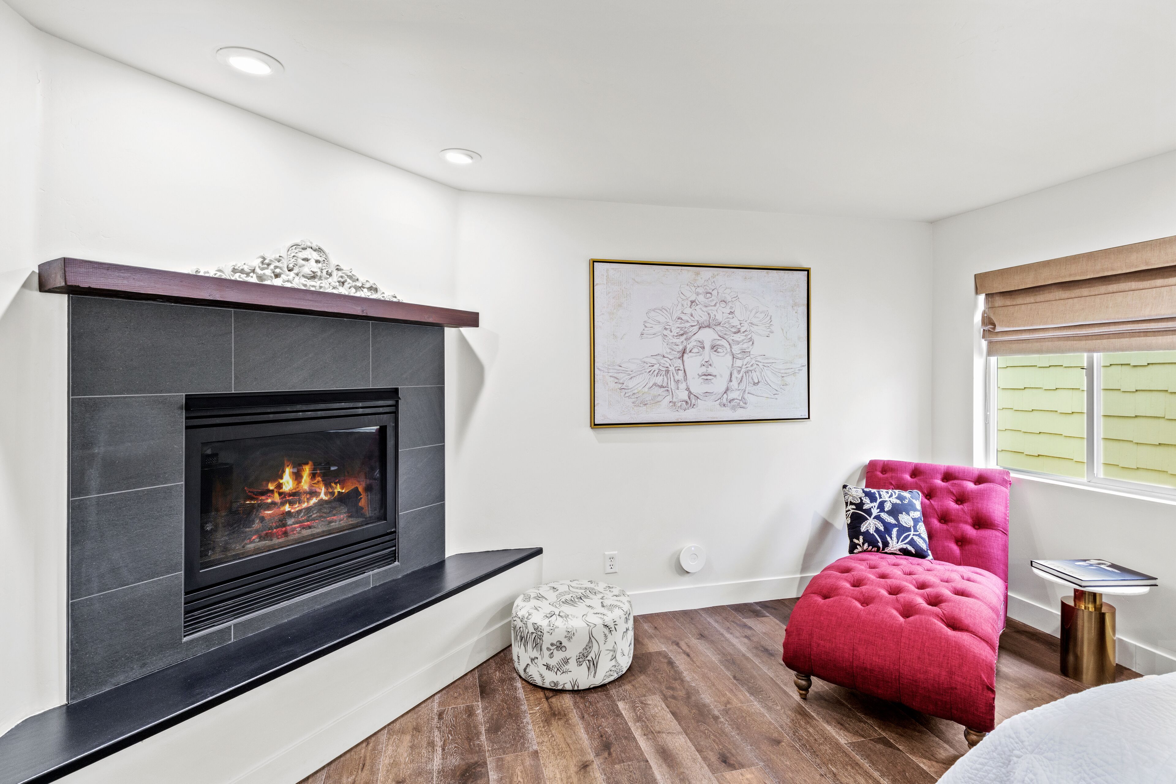 A modern living room with a fireplace, a red ottoman, a small round table, and a framed artwork on the wall.