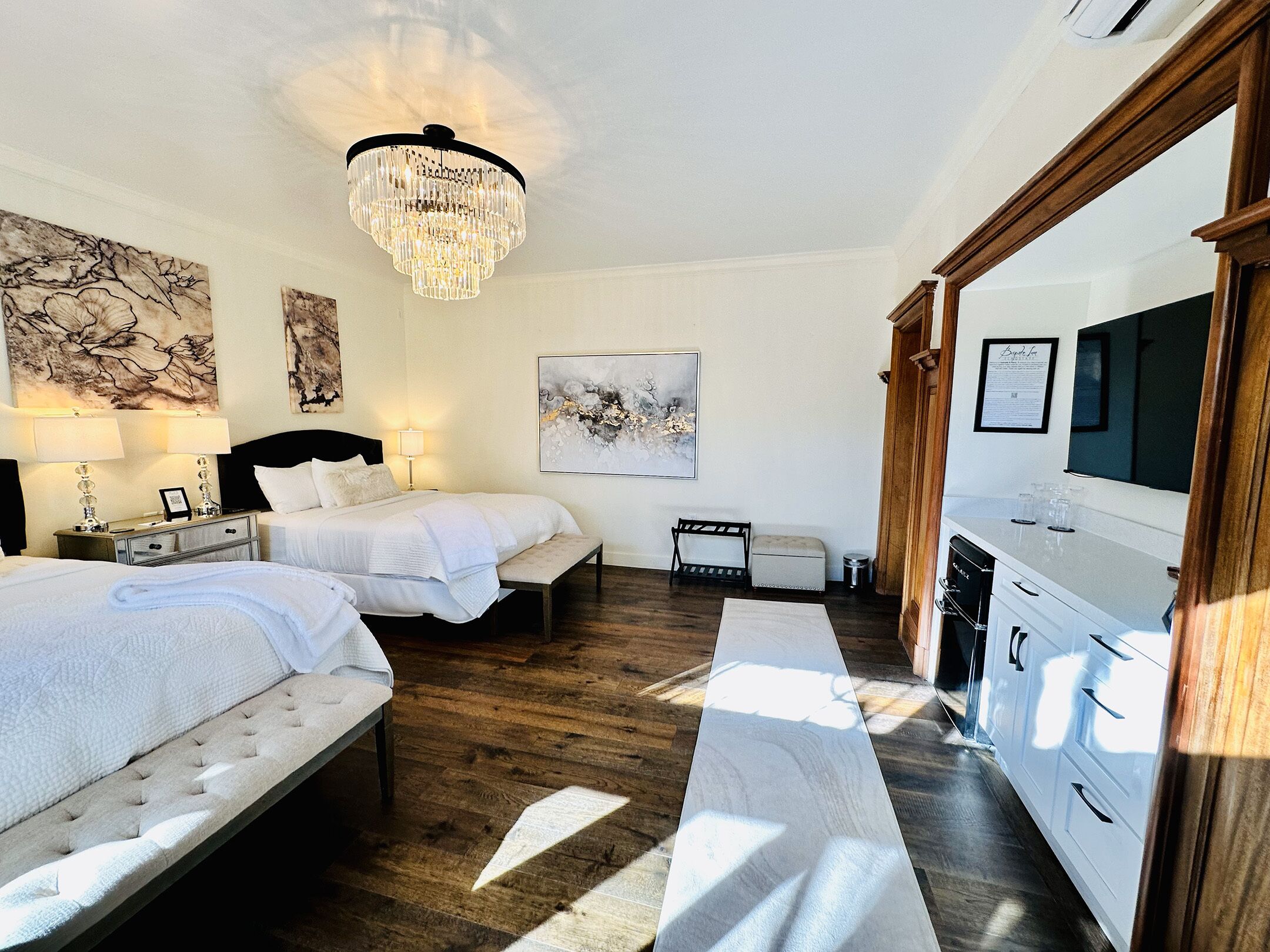 A hotel room with two beds, a chandelier, artwork on the walls, a bench, and a minibar.