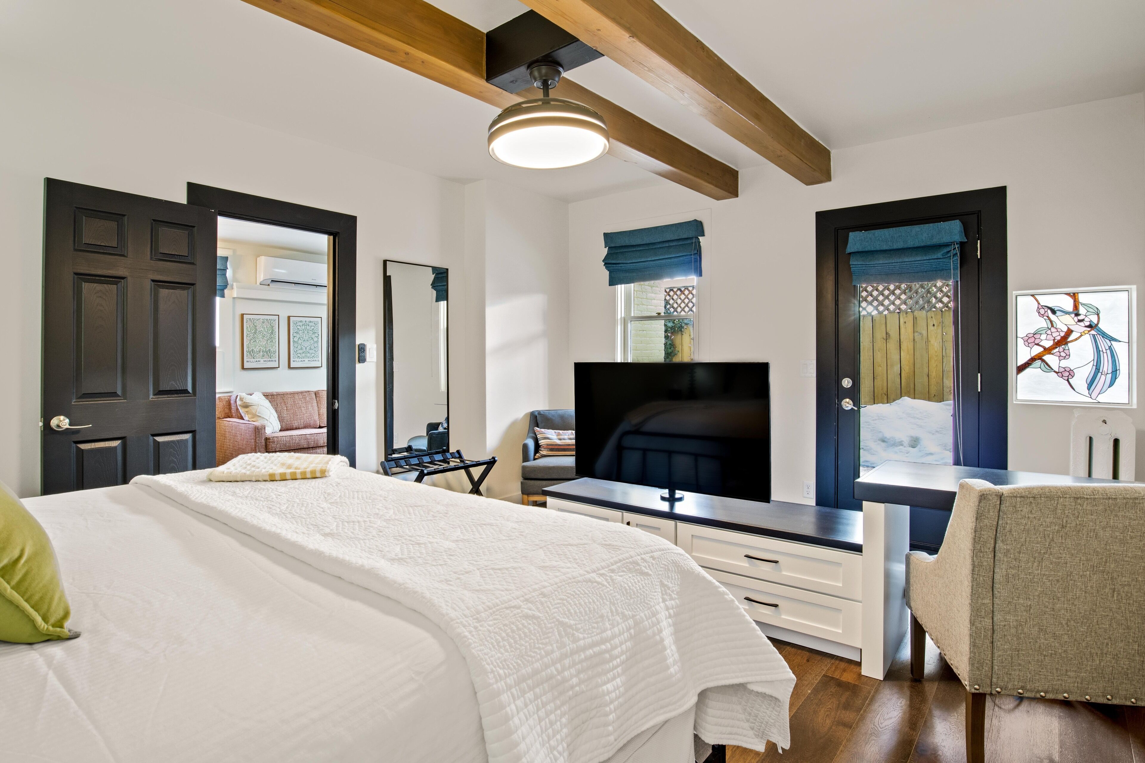 A modern bedroom with a large bed, a TV, a desk, and a chair. There are two doors, one leading to a bathroom and another to a living area.