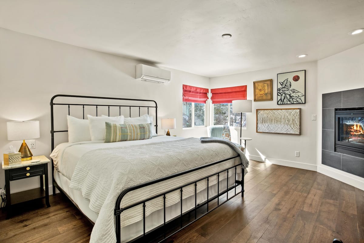 A bedroom with a bed, bedside table, lamp, window with curtains, artwork, and a fireplace.