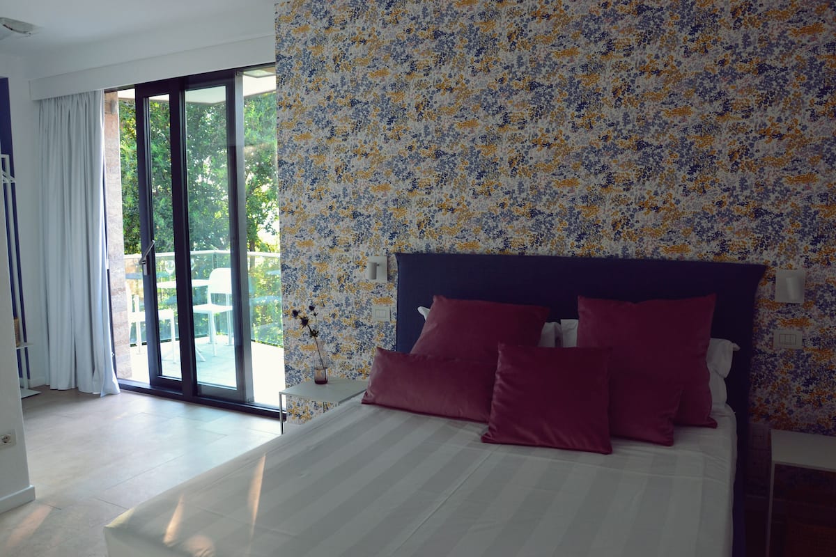 A bedroom with a large bed, a view of the outdoors through a sliding glass door, and a floral-patterned wall.