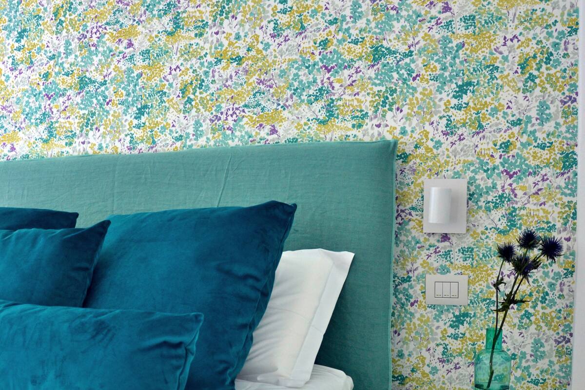 A neatly made bed with teal bedding, a white nightstand with a wicker basket, and a floral-patterned wall.