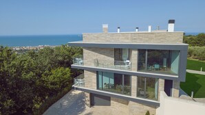 Ett modernt hus i två våningar med stora glasfönster och en balkong, med utsikt över en kuststad och havet.