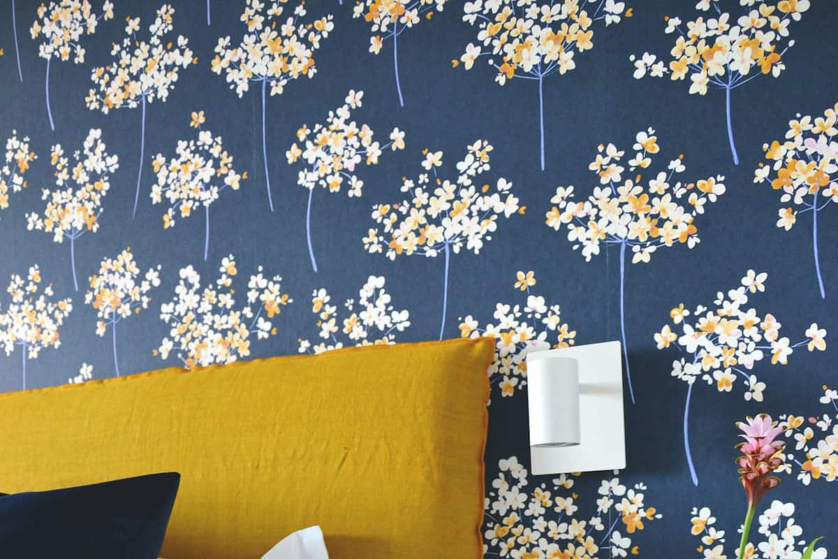 A room with a mustard-colored sofa, a white bedside table, a green vase with a pink flower, and a wall with a floral wallpaper pattern.