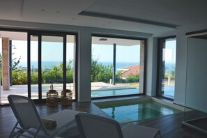 A modern indoor pool area with large windows, a wooden deck, and lounge chairs.