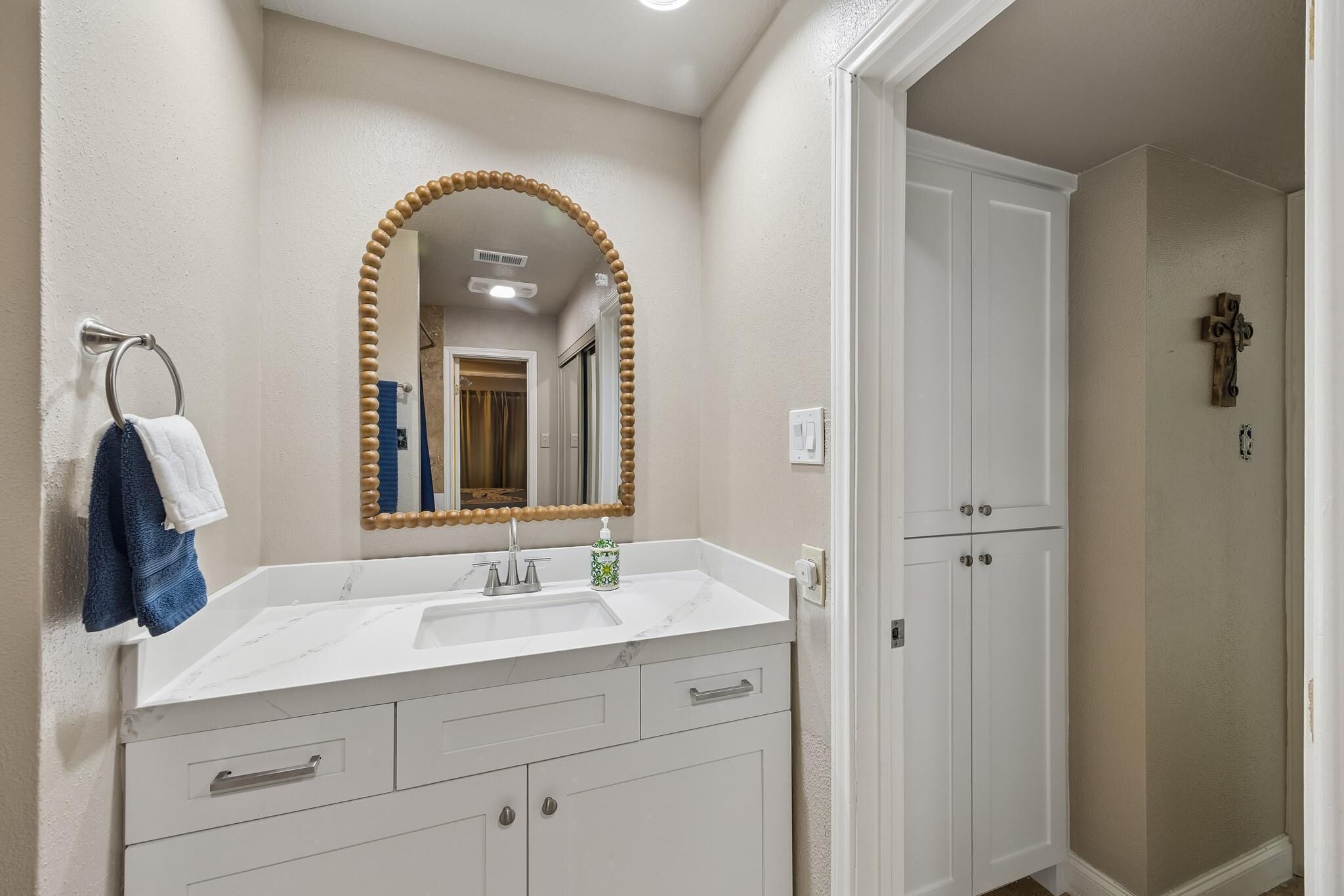 Une salle de bain avec un meuble-lavabo blanc, un miroir avec un cadre en bois et une porte ornée d’une croix sur le mur.