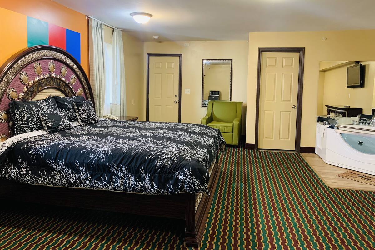 Une chambre d’hôtel avec un grand lit, un fauteuil vert, une salle de bain avec une baignoire et un miroir au mur.