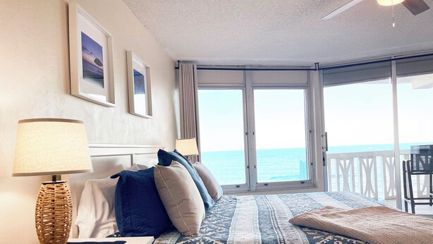 Ein Schlafzimmer mit Bett, Nachttisch, Lampe, einem Fenster mit Meerblick und einem Balkon.