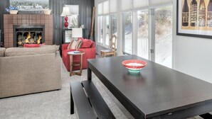 A modern living room with a fireplace, a dining table, and a large window with a view of trees.