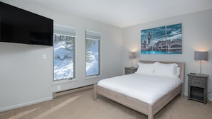 A bedroom with a bed, two bedside tables, a TV mounted on the wall, and a window with a snowy view.
