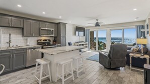 A modern kitchen with grey cabinets, a central island with bar stools, a microwave, and a large window overlooking the ocean. - Ground Floor Gulf Front Oasis On Crystal Beach (Destin)