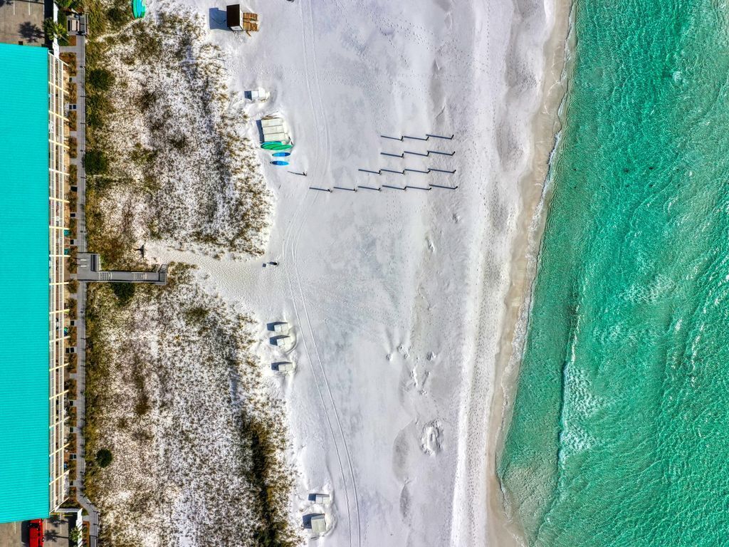 An aerial view of a beach with a clear turquoise sea, a sandy shore, and a few buildings with a simple, modern design.