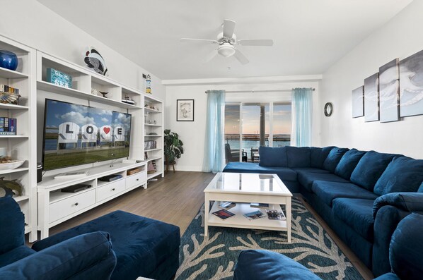 A modern living room with a large flat-screen TV, a white entertainment unit, a blue sofa, a coffee table, and a view of the waterfront through large windows.
