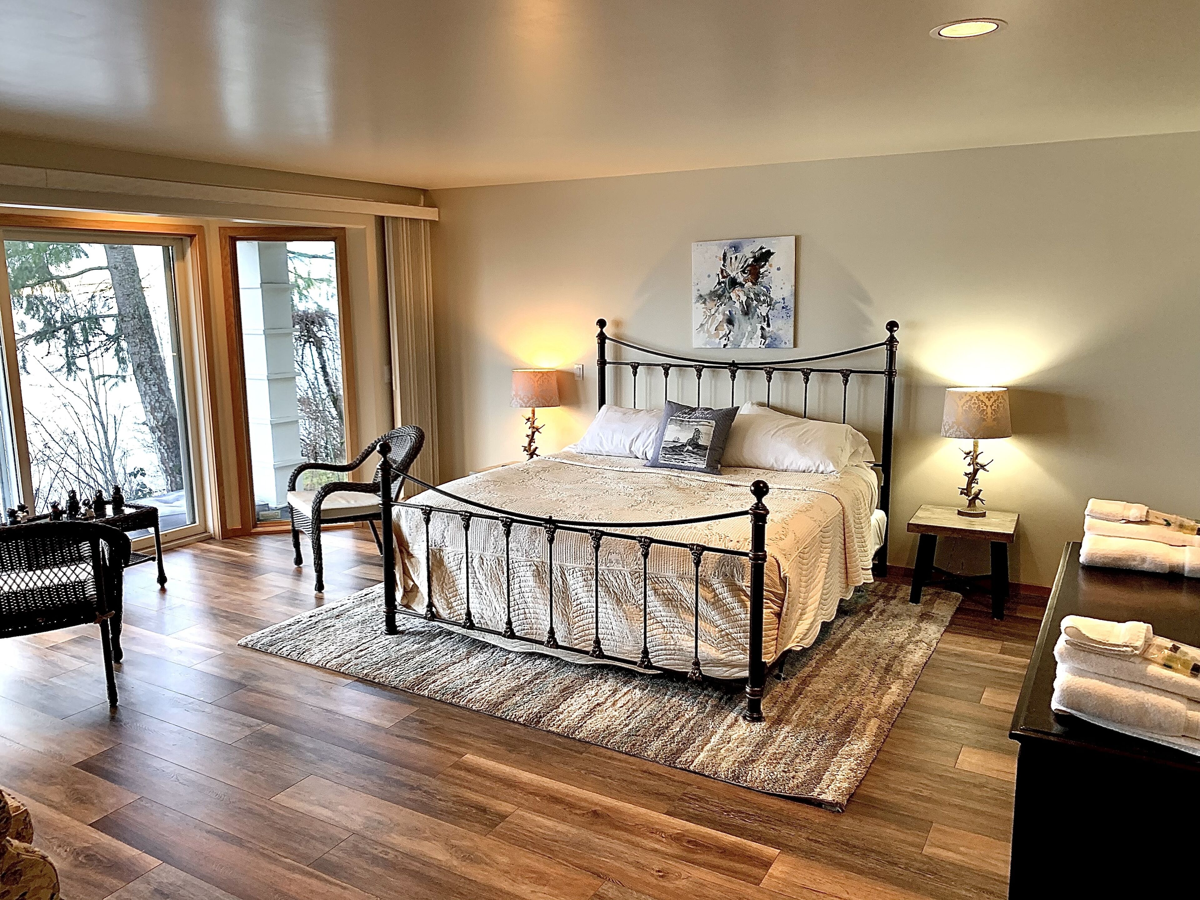 A spacious bedroom with a large bed, two bedside lamps, a chair, and a view of trees through the window.