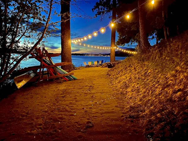A beach path with string lights, a wooden bench, and chairs.