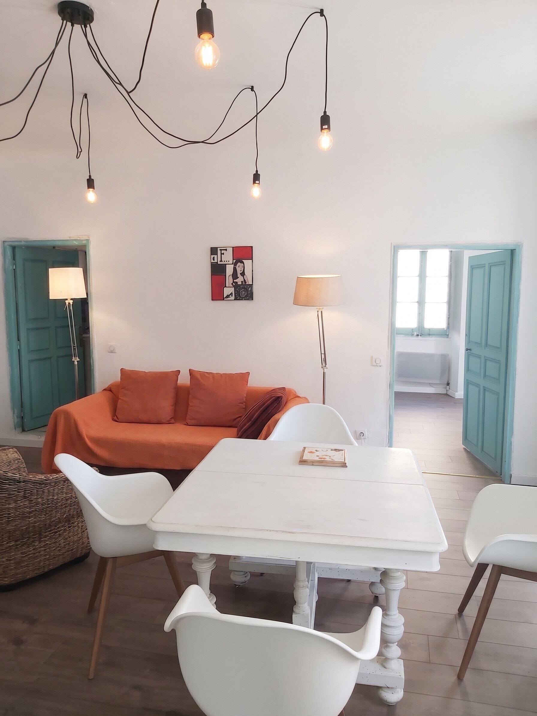 Una stanza con un tavolo bianco, un divano arancione, sedie bianche, una lampada da terra e un lampadario.