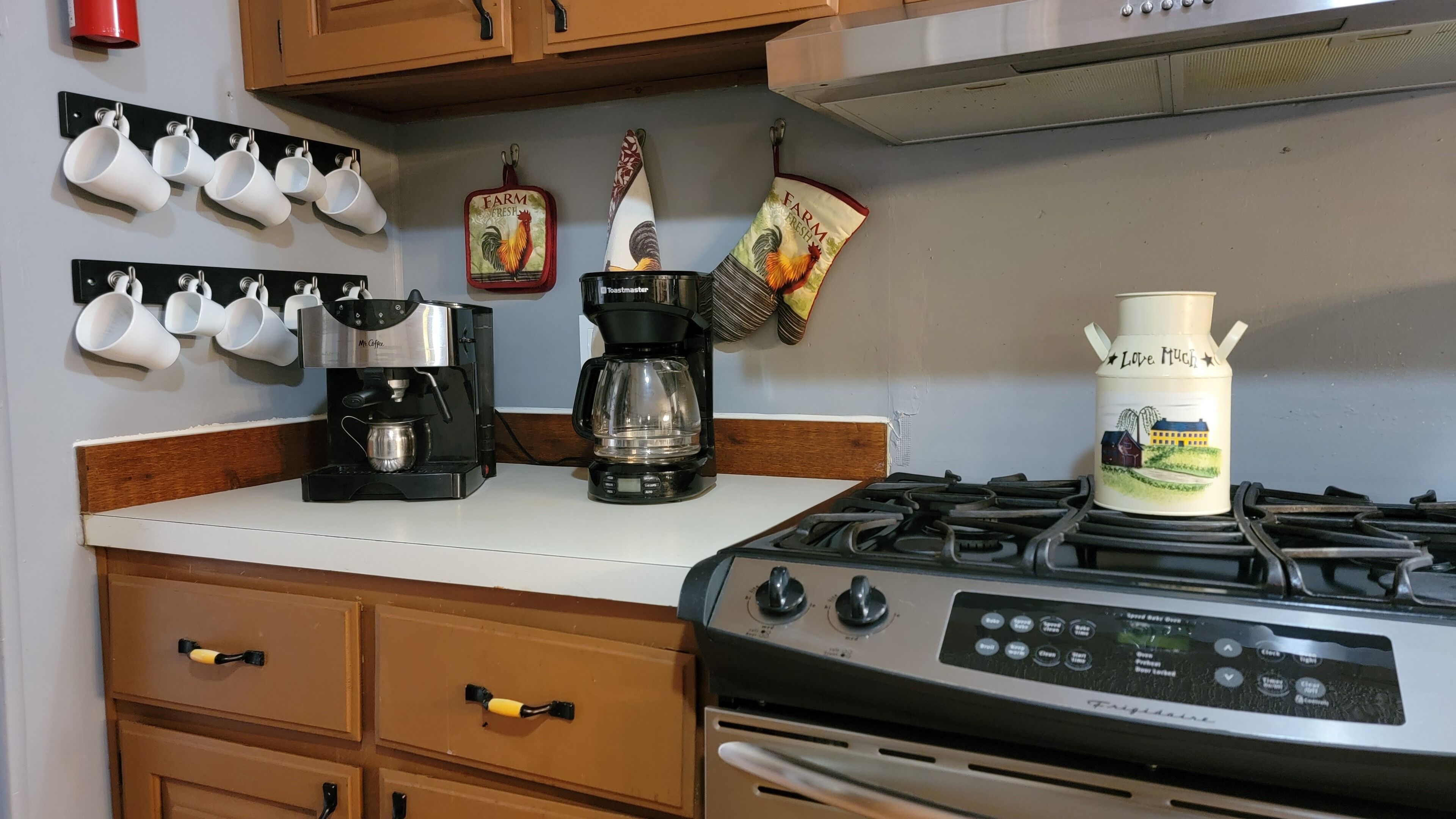 A kitchen with a stove, two coffee makers, and a set of white mugs on a wall-mounted rack.