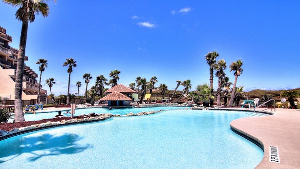 A large swimming pool with a curved design, surrounded by palm trees and a small pavilion. - Easy pool and beach access | MP 114 (Port Aransas)