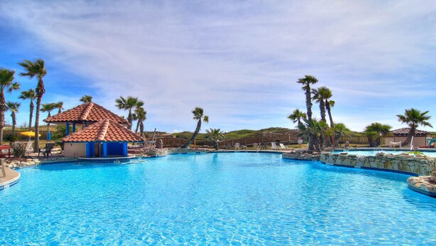A large swimming pool with clear blue water, surrounded by palm trees and a small building with a tiled roof. - Ocean-Pool Views From 2 Balconies | MP315 (Port Aransas)