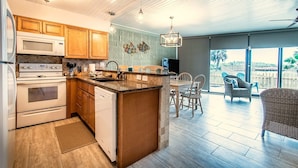 A modern kitchen with wooden cabinets, a built-in microwave, and a dishwasher. There is a dining area with a table and chairs, and a balcony with wicker furniture.