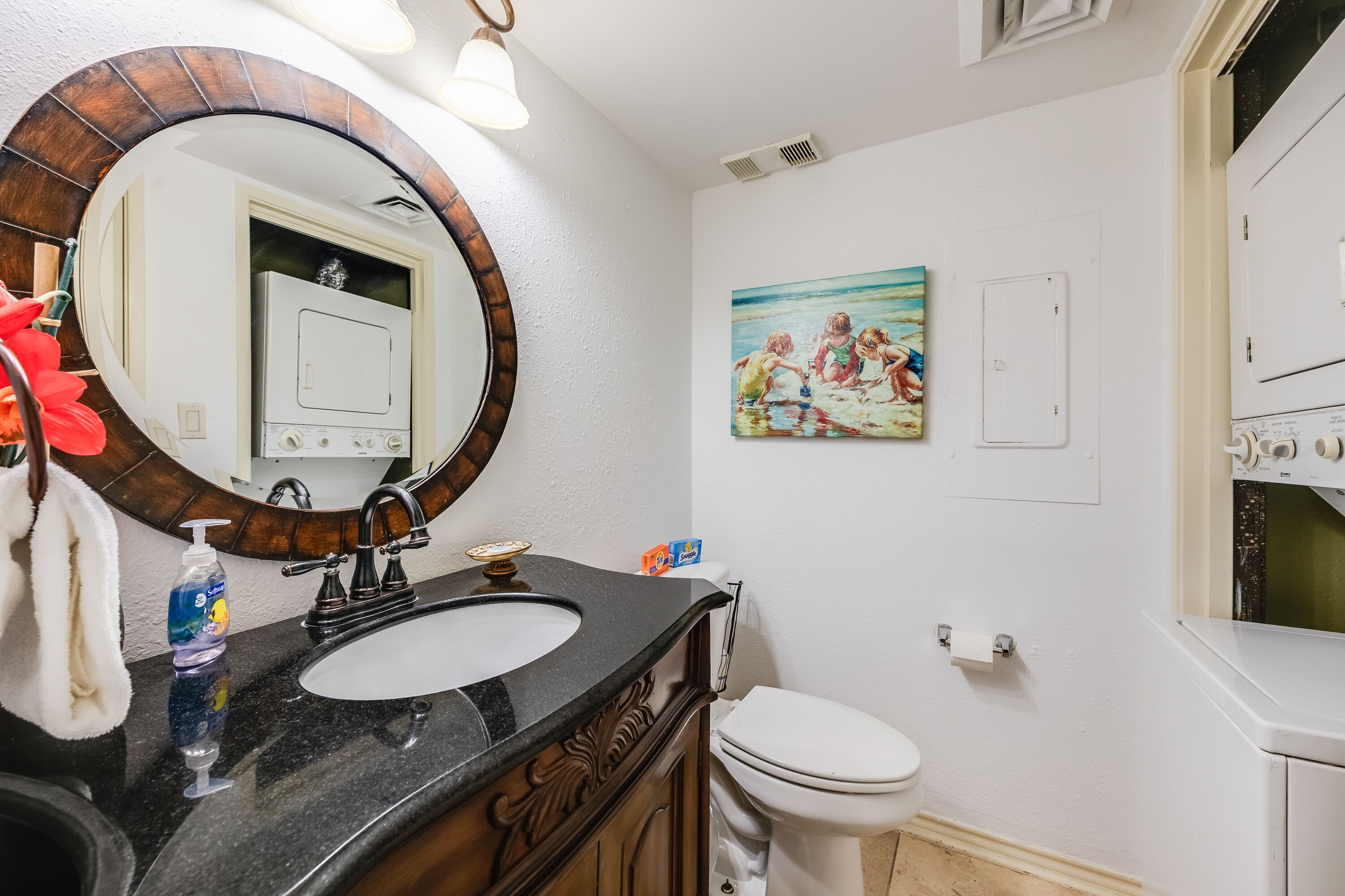 A bathroom with a round mirror, a sink, a toilet, and a painting on the wall.