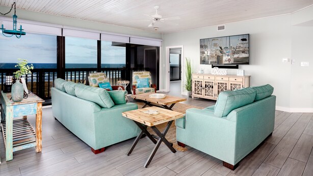 A spacious living area with a large window overlooking the ocean, a ceiling fan, and a television mounted on the wall.