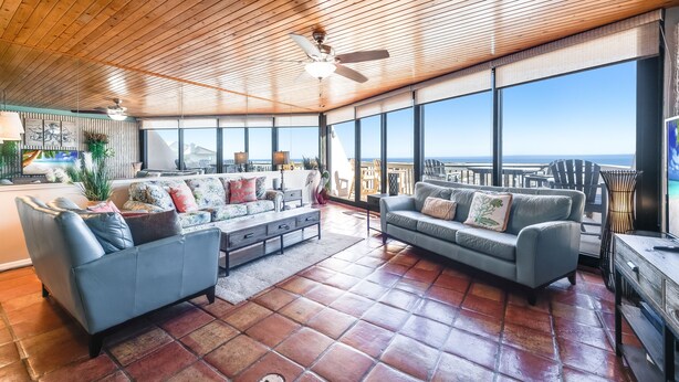 A spacious living area with a high ceiling, large windows offering a view of the ocean, and two sofas with cushions.
