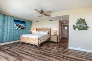 Quarto com cama de madeira, ventilador de teto, mesa de cabeceira com abajur e um peixe decorativo na parede.