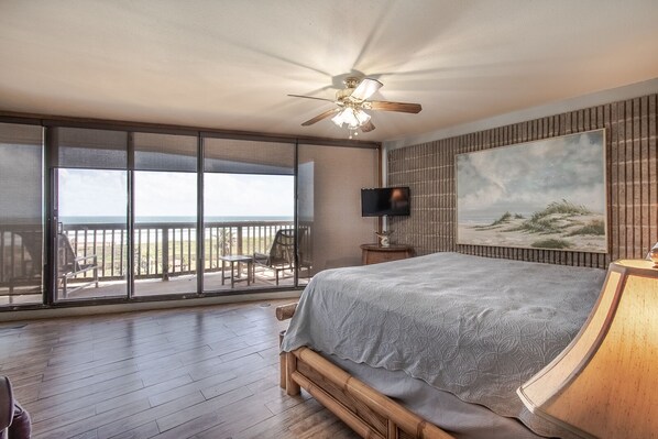 A bedroom with a large bed, a ceiling fan, a television, and a painting of a beach scene.