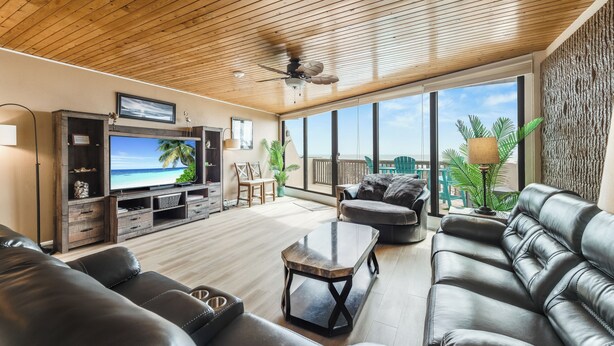 Una sala de estar amplia con techo de madera, un televisor de pantalla plana grande, sofás de cuero y vista al océano a través de ventanas amplias.