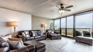A spacious living room with a large sectional sofa, a chaise lounge, and a small ottoman. There is a ceiling fan, a large window with a view of the outdoors, and a wooden deck visible through the glass doors.
