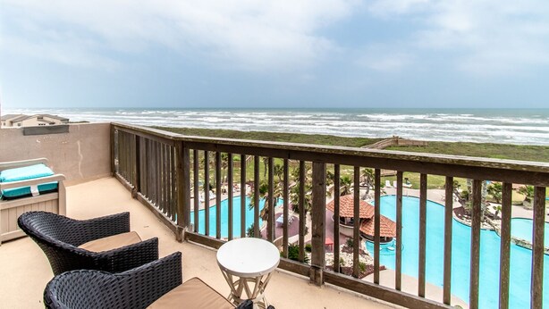 A balcony with wicker chairs and a small table, overlooking a swimming pool and a view of the ocean.