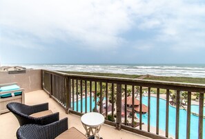 A balcony with wicker chairs and a small table, overlooking a swimming pool and a view of the ocean. - Port A Paradise | Your Family's Beach Destination (Port Aransas)