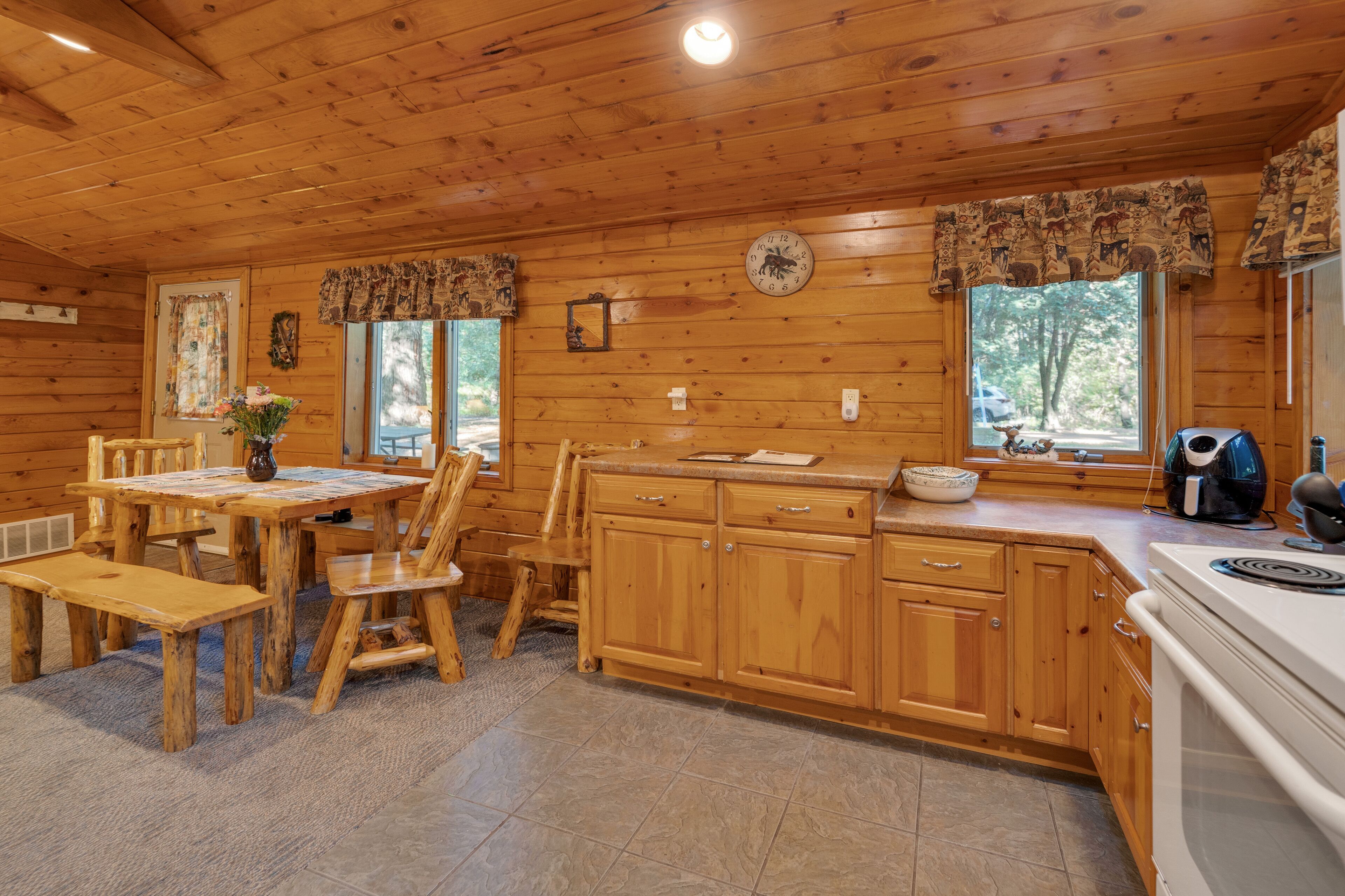 Interior de una cabaña de madera con una mesa de comedor, sillas, armarios de madera, una encimera de cocina y un ventanal grande.