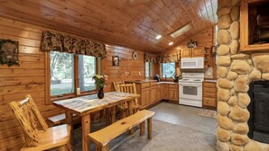 A wooden cabin interior with a dining table, chairs, a stone fireplace, a kitchen with a microwave, and a clock on the wall. - Wisc Dells Amish Cabin in Woods for 10 people (Wisconsin Dells)