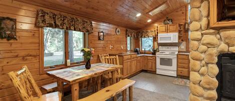 A wooden cabin interior with a dining table, chairs, a stone fireplace, a kitchen with a microwave, and a clock on the wall.
