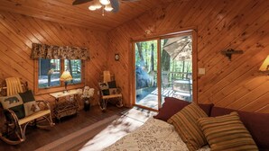 A cozy cabin interior with a wooden ceiling, a rocking chair, a desk with a lamp, a bed with patterned bedding, and a sliding glass door leading to an outdoor deck. - Wisc Dells Amish Cabin in Woods for 10 people (Wisconsin Dells)