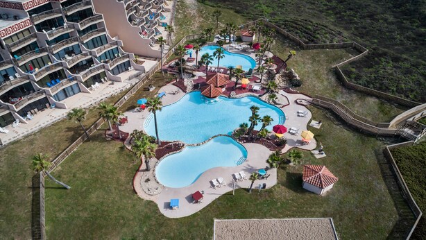 An aerial view of a resort with multiple swimming pools, palm trees, and a large building with balconies. - Sassy Seagull @ The Mayan Princess (Port Aransas)