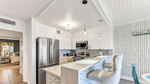 A modern kitchen with a stainless steel refrigerator, a microwave, and a sink. There is a bar area with a white marble countertop and a bar stool. The kitchen has white cabinetry and a patterned backsplash. - Poolside views | Private access to beach | Beachfront | MP103 (Port Aransas)