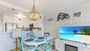 A modern kitchen with white cabinets, a dining area with blue chairs, and a TV displaying a beach scene. - Poolside views | Private access to beach | Beachfront | MP103 (Port Aransas)