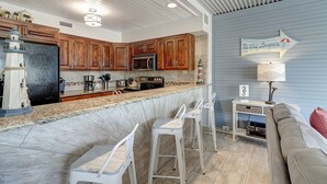 A kitchen with wooden cabinets, a granite countertop, and a bar area with white stools. - A Beachy Dream MP118 | Resort Pools | Private Boardwalk (Port Aransas)