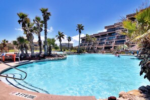 A swimming pool with a 3 ft no diving sign, surrounded by palm trees and a multi-story building with balconies.