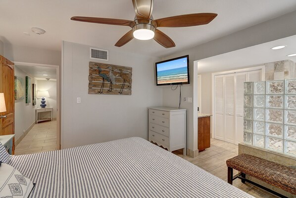 A bedroom with a bed, a ceiling fan, a television, a dresser, a bench, and a view of the beach through a window.