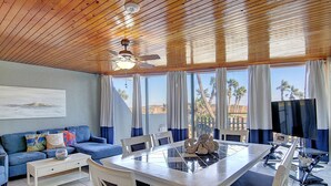 A room with a wooden ceiling, a dining table with chairs, a sofa, a TV, and large windows with curtains. - Beachfront Vibes @ MP109 | Poolside Views | Beach Access (Port Aransas)