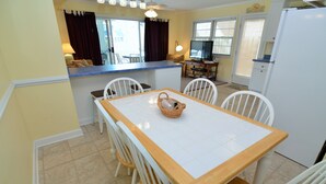 Un comedor con mesa de madera y sillas blancas, una cocina con refrigerador y un televisor.