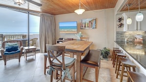 A dining area with a wooden table and chairs, a view of the ocean through large windows, and a kitchen with a bar counter.